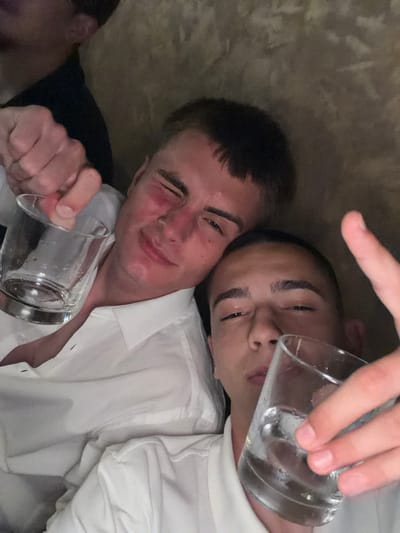 The image displays a close-up, portrait-oriented view of two young adult males in an indoor setting. No discernible visual information within the image provides any indication of the city of Slivnitsa, Bulgaria, or any specific geographic location.
The male positioned in the upper portion of the frame has short dark hair and fair skin. He is wearing a white collared shirt and is seen winking or squinting his right eye, with a slight smile on his face. His cheeks and nose appear flushed with a reddish tone. He holds a clear, short drinking glass containing a clear liquid and ice cubes in his right hand, which is clenched around the upper portion of the glass.
The second male, positioned in the lower portion of the frame, has very short dark hair and a slightly darker complexion. He is also wearing a white collared shirt. He looks directly towards the viewer with slightly narrowed eyes and a neutral expression. He holds a similar clear drinking glass, also containing clear liquid and ice cubes, in his right hand, with fingers wrapped around the glass.
The two individuals are positioned closely together, suggesting an intimate or friendly interaction. The background is indistinct, appearing as a mottled brown or tan surface, possibly an upholstered wall or furniture, indicating an indoor environment. The lighting is dim and warm, contributing to a slightly soft-focus or grainy appearance in some areas of the image. A third person's indistinct head and shoulder are partially visible in the far upper left corner.