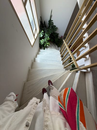 View of the interior of a building in Glifada, Greece, with a woman descending a spiral staircase. The staircase is made of white marble. A wooden pergola-like structure is above the stairs. The woman is wearing beige pants, a white top with lace trim, and burgundy shoes. She also has a colorful striped shoulder bag. On the landing, there is a window and a potted plant. A broom is leaning against the pergola.