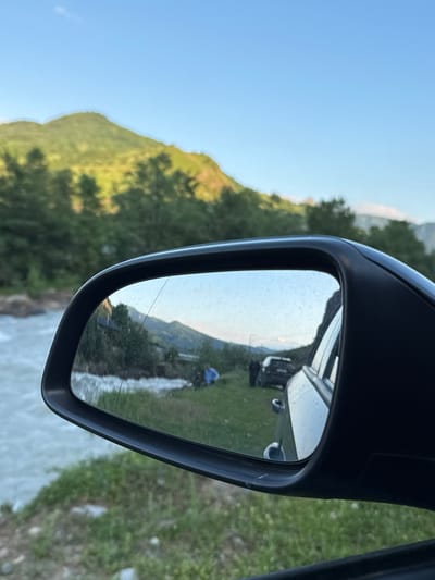 The image shows a view from inside a car, looking at the side mirror, which reflects a scenic landscape. The primary focus is the reflection in the side mirror, which reveals a body of water, possibly a river, flowing to the left of the frame. Beyond the water, there is a grassy embankment with some trees. In the reflection, two figures are visible on the grassy area. One figure, wearing a blue top, is standing near the water's edge. Further back, two dark-colored vehicles are parked. The background of the reflection shows a lush green mountainside under a clear blue sky, with some trees in the middle ground. Outside the mirror, the real-world view shows the side of the car, the grassy ground, and a glimpse of the river to the left. The overall scene suggests an outdoor setting in a rural or natural environment.
