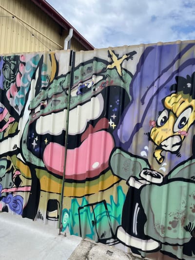 A vibrant, cartoonish graffiti mural is depicted on a corrugated, possibly translucent, metal surface. The image is oriented with the sky on the left, suggesting a rotation from its original upright position.

The mural features an array of brightly colored, stylized figures and abstract shapes. In the upper part of the artwork (as oriented in the image), a yellow, wide-eyed creature with a whimsical expression looks upwards. Below it and to the left, a green, blob-like entity with an eye and a yellow star-shaped object above it is visible. Dominating the center-right is a large, glossy pink, pill-shaped or bubble-gum-like form with a white highlight, set against a dark background speckled with small white and yellow stars. Other parts of the mural include various green, purple, black, and white abstract shapes and outlines, contributing to a dynamic and playful composition.

To the left of the graffiti (which would be above it in a standard orientation), a bright blue sky with scattered white clouds is visible, indicating daytime. Below the sky, architectural elements include a yellow-sided building with horizontal panels, a brown roofline, and a white downspout. To the far right of the mural, a grey concrete surface is partially visible. The overall impression is one of energetic and creative street art. No discernible text is visible within the artwork or the surrounding environment.