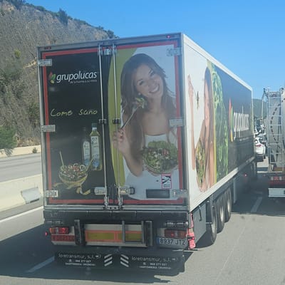 The image shows the rear of a large delivery truck, photographed from a slightly elevated perspective. The truck's rear doors are decorated with a food-related advertisement featuring a woman eating a salad and the brand "Grupolucas" with the tagline "de la huerta a su mesa." The ad also includes the phrase "Come Sano," along with depictions of olive oil and another salad tied with measuring tape. A small "Attention Angles Morts" sticker is visible. The license plate number "8937 JTZ" and the contact information for "Loretransmur, S.L." of Santomera (Murcia) are also displayed. The truck is traveling along a road. A mountainous, green landscape is visible in the background.