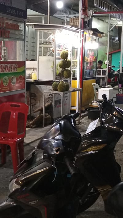 This image captures a street-side durian stall at night in Bandarbireuen, Indonesia. The primary subjects are several durians displayed on a metal rack, with more durians piled on the ground and inside a cooler. A red plastic chair sits in the foreground to the left, and a black motorcycle is parked in front of the stall, partially obscuring the view.
In the background, a young man, likely a vendor, is visible inside the stall. The stall itself is a simple structure with a metal frame and transparent panels, illuminated by bright lights. Behind the durian stall, another vendor stall selling various packaged goods is visible, with bright signage.
The scene suggests a typical evening at a local food stall in Indonesia, where durians are a popular seasonal fruit. The lighting indicates it is nighttime, and the overall atmosphere is lively and commercial.
Visible text includes "LEVEL 1-10" on a sign at the top left, and "REK KAK EVI 2" on a colorful advertisement to the left of the durian stall. The word "ajim" is visible on the cooler. The text on the vendor stall in the background is difficult to read but appears to be in Indonesian.
