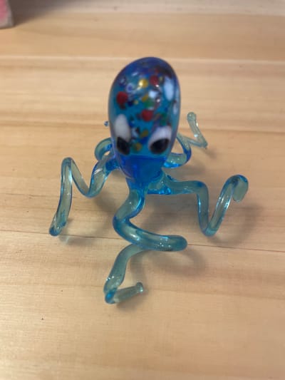 A small, translucent blue glass octopus figurine with colorful flecks and googly eyes rests on a light-colored wooden surface. The octopus has eight curled tentacles spread out around its body. The surface appears to be a wooden tabletop or floor. The image is a close-up shot, with the figurine centrally located. The background is out of focus. There is no indication of the city of Taganrog, Russia, or any specific location within a city.