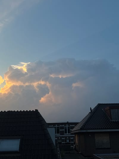A vertical, eye-level shot captures a dramatic sky over the rooftops of Putten, Netherlands. The upper portion of the image is dominated by a vast expanse of blue sky, with subtle linear cloud formations visible in the upper right quadrant. A massive, cumulonimbus cloud formation occupies the center and right of the frame, its thick, white mass dramatically illuminated by the warm glow of the setting sun from the left. The light creates striking contrasts, highlighting the textured contours of the clouds.
In the foreground, two dark, tiled rooftops are visible. The left roof, closer to the viewer, features a light-colored dormer window with horizontal blinds. To its right, a building with a dark tiled roof and brick facade is partially visible. This building's windows are boarded up with plywood, suggesting a vacant or under-construction property. A lone black bird is perched on the peak of this roof. The overall scene depicts an urban residential area under a dynamic, illuminated sky.
