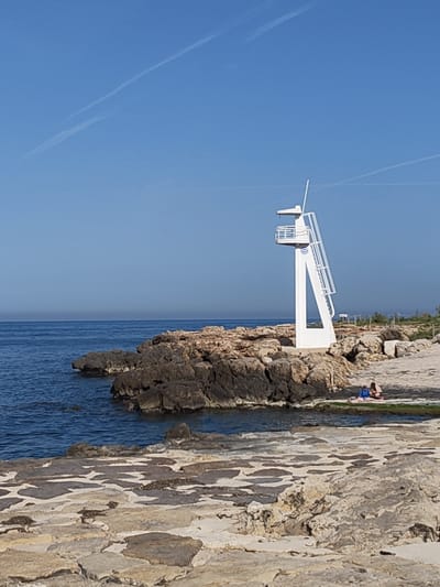 The image depicts a coastal scene in Rotes, Spain. A white lifeguard tower stands prominently on a rocky outcrop jutting into the blue sea under a clear sky. A small group of people are seated on a blanket near the base of the rocks. The foreground consists of flat, interlocking stone slabs. The atmosphere is bright and sunny.