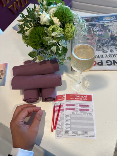 An indoor scene on a white tabletop depicts a hospitality setting at Epsom Downs Racecourse in Epsom and Ewell, United Kingdom, likely during a horse racing event. A clear champagne flute, filled with a sparkling yellow beverage and bubbles, stands prominently. To its left, a small floral arrangement in a reflective silver vase features white roses, green hydrangeas, spiky green Dianthus 'Green Trick', and variegated ivy. Four rolled, deep purple fabric napkins are stacked horizontally in the center of the table. To the right, two white paper forms titled "placepot and multi-leg bets," with red headers, are laid out; one is fully visible, detailing betting options for six races across categories like "placepot," "quadpot," "jackpot," and "scoop6." Two red pens rest on the partially visible betting slip to the left. A folded newspaper, partially obscured behind the champagne glass, shows headlines related to horse racing, including "DERBY REL" and "2ND? PAID AS WINNER," along with the partial name "Betfred." The lower part of a person's right hand, wearing a white shirt cuff, rests on the table in the lower left corner. The background reveals blurred purple and white elements, possibly seating or decor.