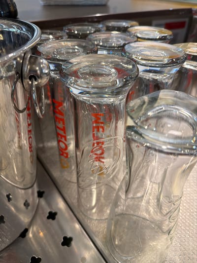 The image shows a close-up, overhead view of several clear glass beer mugs with red "METEOR" branding. The mugs are stacked and arranged on a metallic surface. Some of the mugs are upside down, showing their bases, while others are right-side up. The metallic surface has small, decorative cutouts resembling four-leaf clovers. In the background, a blurred metallic bucket or container with handles is partially visible. The scene appears to be in a bar or restaurant setting in Vienne, France, possibly within a brewery or establishment that serves Meteor beer, given the prominent branding on the glasses. The lighting suggests an indoor environment. There are no people visible in the image.
