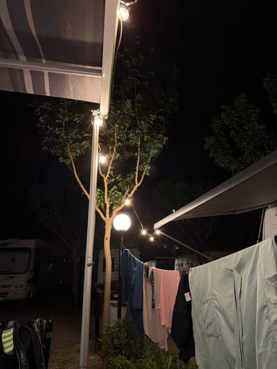 The image depicts an outdoor scene at night, likely at a campsite in or near Πόρτες, Greece. In the foreground, laundry is hanging on a clothesline. The laundry includes items of various colors and textures, such as light blue, pink, black, and a patterned garment. To the left of the clothesline, a white camper van is partially visible.

In the mid-ground, a tree with bare branches and sparse leaves is illuminated by a round, white lamp at its base. String lights with visible bulbs are also strung along a pole and a structure that appears to be an awning. This awning, with its striped fabric, extends over the camper van. Another awning, made of a darker, solid material, is visible in the background, also over a camper van.

The background is dark, with the silhouettes of trees and foliage against the night sky. The overall impression is one of a temporary campsite set up for the night, with elements of comfort and domesticity (laundry, lighting) juxtaposed with the natural environment. There are no people visible in the frame.