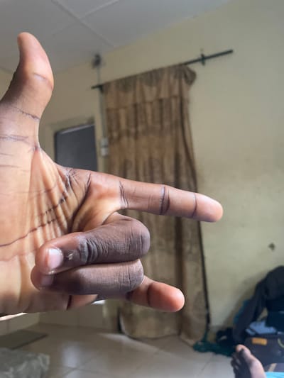 The image shows a close-up of a dark-skinned hand making a gesture, likely the "I love you" sign, with the thumb and index finger extended and the other fingers curled into the palm. The hand is positioned in the foreground, dominating the frame. In the background, a lightly colored wall is visible, with a dark curtain hanging from a rod. To the right, a cluttered area shows piles of clothes and what appears to be a shoe or bag. The floor is tiled. The overall environment suggests an indoor residential setting, possibly in Malete, Nigeria. The lighting appears to be natural, coming from an unseen source.