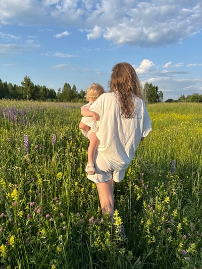 A person, viewed from behind, stands in a vibrant field of wildflowers under a blue sky with scattered white clouds. The person is holding a small child, also facing away from the camera. The person is wearing a white t-shirt and light-colored shorts, and the child is wearing a white dress. The field is filled with various wildflowers, including purple lupine and yellow flowers, with a line of trees in the distance. The scene suggests a peaceful, natural setting, possibly during a warm day. The location is identified as the city of Koloksha, Russia.