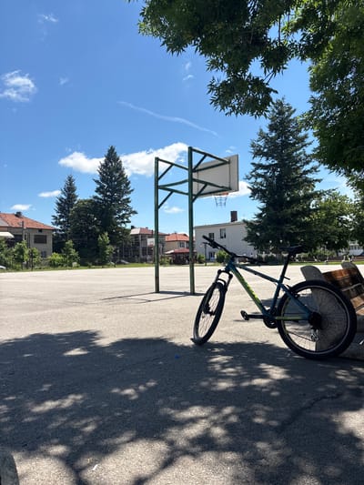 The image depicts an outdoor basketball court in Bozhurishte, Bulgaria, on a sunny day with a clear blue sky and scattered clouds. A bicycle is parked near a basketball hoop, leaning against a wooden bench. The court is paved with asphalt, and the shadows of trees are cast across its surface. In the background, there are several residential buildings and tall evergreen trees. The scene appears to be a quiet, public recreational area.