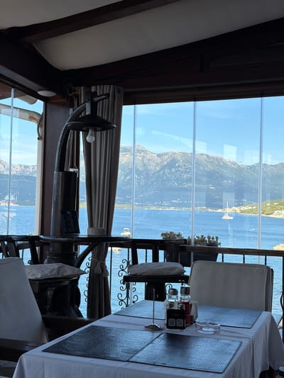 The image shows an interior view of a restaurant in Krašići, Montenegro, overlooking the Bay of Kotor. A large, vertically-oriented window offers a view of the mountains, the sea, and the town. Tables with white tablecloths and reptile print placemats are set for dining. There are chairs with white cushions. A decorative helm mounted above the window adds a nautical theme to the space. Table accessories such as condiment holders and QR code stands are present on the tables.