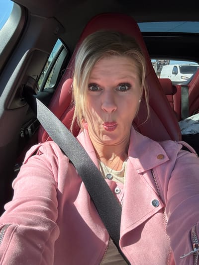 A blonde woman is depicted in a brightly lit car interior, likely taking a selfie or being photographed from an angled perspective. She is seated on a rich red leather car seat, wearing a light pink jacket with silver snap buttons and zippers, over a lighter-colored top adorned with a delicate necklace featuring a heart-shaped charm. A dark seatbelt diagonally crosses her chest and shoulder.
The woman has light-colored eyes and her lips are slightly pursed in a posed expression, perhaps playful or coquettish. Strong sunlight illuminates the scene, casting distinct shadows across her face and clothing, indicating a sunny day. Through the car window, a partial view of another white vehicle, possibly a truck or trailer, and some indistinct outdoor elements are visible. The car's interior also features dark-colored trim elements. The image is rotated counter-clockwise, adding to its candid, dynamic feel. The inferred location is Bredene, Belgium. No text is visible in the image.