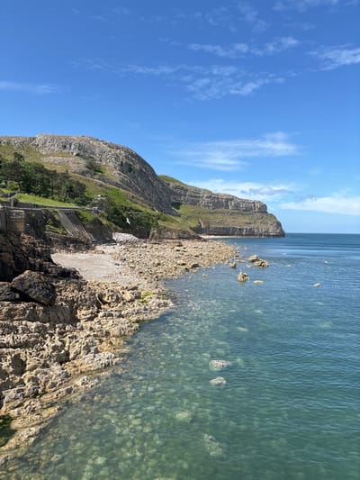 The image captures a coastal scene in Llandudno, United Kingdom. A rocky shoreline transitions into clear, shallow water with visible pebbles at the bottom. Adjacent to the water is a cliff face, with vegetation and a set of stone steps visible. The sky above is blue with wispy clouds.