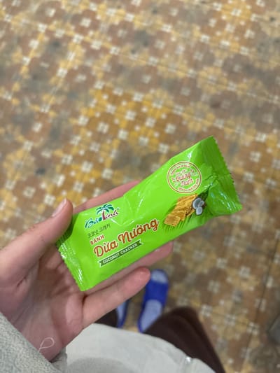 The scene is documented in Nam Hải Lăng Commune, Vietnam. A fair-skinned left hand occupies the central and lower-left portion of the frame, holding a bright green foil package horizontally. The hand's fingers wrap around the package, with the thumb positioned on top. The lower-left edge of a light grey, possibly fabric or towel, material is visible beneath the hand.
The package is a "Bảo Linh" brand "Coconut Cracker" ("BÁNH DỪA NƯỚNG" in Vietnamese and "코코넛 크래커" in Korean). The front of the package features a stylized palm tree logo, product text in white and yellow, and an illustration of light brown rectangular crackers alongside two halves of a coconut with white flesh. A circular green seal or sticker on the upper right of the package contains Vietnamese text, partially legible as "Quảng Nam," indicating a product origin or association with Quảng Nam province.
The background consists of a tiled floor exhibiting a repeating mosaic pattern composed of small squares in shades of brown, beige, and yellow. Below the package and hand, blurred in the mid-ground, a pair of blue open-toed sandals or slippers is visible, with a dark brown fabric (likely clothing or part of a leg) partially obscuring one of the sandals.