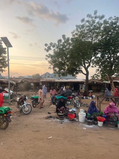 This image captures an active street scene in Rinjim Gani, Nigeria, during late afternoon or early evening. The sky displays a pale orange-yellow hue with scattered light clouds, indicating a sunset.
The ground is unpaved and dusty, consistent with a rural or semi-urban environment. Numerous single-story structures with corrugated metal roofs are visible in the background, suggesting dwellings or small businesses. A large deciduous tree with sparse foliage stands prominently on the right side of the frame, and another smaller tree is on the far left. Overhead power lines traverse the upper middle section of the image. A solar-powered street light or panel is affixed to a pole on the far left.
Several individuals, predominantly women, are engaged in activities on the ground. Three women in the foreground are seated directly on the dirt, each operating small charcoal stoves with pots and basins, indicating food preparation or vending. One woman on the far left wears a dark head covering and clothing, stirring contents in a pot. Another woman in the mid-right wears a blue head covering and garment, attending to multiple containers. A third woman in a pink head covering and garment sits further right, also with cooking implements. Various foodstuffs are visible in red plastic bowls and metal pots. White plastic buckets are interspersed among their setups.
Multiple motorcycles, commonly used for transport, are present. A silver motorcycle is parked in the mid-left, a darker one is positioned to its right, and the rear of a red motorcycle with a green compartment is visible on the far left.
Other individuals are standing or sitting. A man in a light brown traditional garment stands near the silver motorcycle, looking towards the right. A group of men, one in a plaid shirt, are visible sitting or standing on the far left, near the red motorcycle. In the background, other figures, some draped in light blue or red head coverings, are seen walking or standing near the structures. A rudimentary wooden table or stand is present on the far right. The scene depicts a bustling local gathering point with ongoing commercial and social activity.