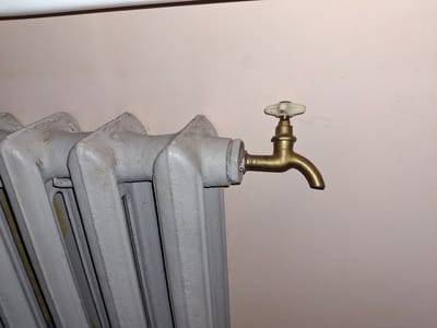 This media file is a close-up photograph of a radiator with an attached brass valve. The radiator is painted light gray and appears to be made of cast iron, with multiple vertical sections. The valve has a curved brass spout and a white ceramic handle. The background is a plain, light-colored wall, possibly painted. There are no people or other objects clearly visible. The lighting suggests an indoor setting, possibly a room in a home or building. The image focuses on the functional components of the heating system. No text is visible in the image. The location context provided is Ogre, Latvia.