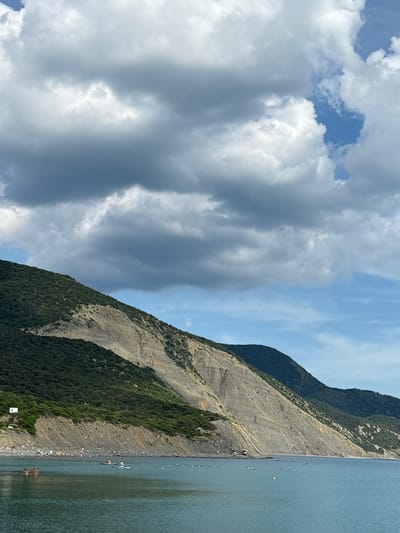 The image depicts a coastal landscape in Bol'shoi Utrish, Russia, under a partly cloudy sky. The upper half of the frame is dominated by a dense formation of grey and white cumulus clouds, with patches of clear blue sky visible in the upper right and center.

The middle and lower portions of the image feature a prominent mountainous coastline. The mountains exhibit steep slopes covered in dense green vegetation, with significant sections of exposed light-colored rock, likely due to geological strata or erosion. The coastline curves gently from left to right.

At the base of the mountains, a narrow, pebbly beach is visible. On this beach, numerous small figures representing people are dispersed, suggesting recreational activity. Some colorful accents, possibly beach equipment or small shelters, are also discernible on the sand. A small, white rectangular structure is perched on the green hillside on the far left.

The foreground is occupied by calm, dark blue-green water. A line of white spherical buoys extends horizontally across the water from the left, marking an area in the sea. Further out, a single red conical buoy is visible. Two individuals are positioned on separate stand-up paddleboards, traversing the water in the mid-ground. Closer to the shore on the left, two individuals are seated on a low-profile, square-shaped floating platform or raft. The scene suggests a popular coastal area during daylight hours.