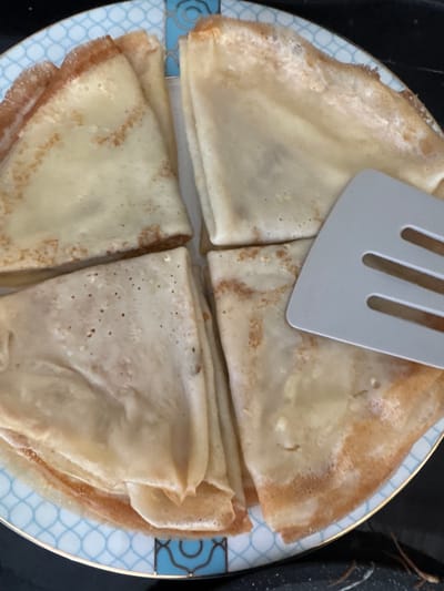 A top-down close-up view shows four folded crepes on a plate with a blue and white pattern. A light gray spatula with three horizontal slots is placed on top of the crepes. The crepes are golden brown with some darker, crispier edges. Some crepes appear slightly filled, showing a darker color inside. The plate has a thin gold rim. The background is dark and out of focus. The scene is set indoors, likely in a kitchen or dining area. There is no information to indicate the location within the city of Uyutnoye, Ukraine.