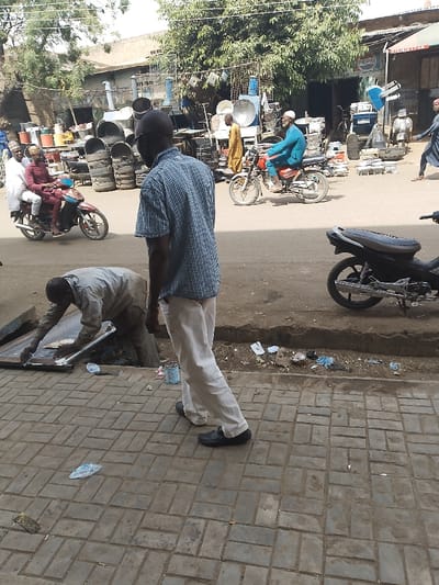 The scene depicts a bustling street in Kofar Mazugal, Nigeria. In the foreground, a dark-skinned adult male is captured from behind, wearing a blue and white checkered short-sleeved shirt, light-colored trousers, and dark shoes, walking away from the viewer on a patterned brick paver sidewalk. To his left and slightly forward, another dark-skinned adult male, dressed in light gray work attire, is bent over an open metal access panel or grate on the sidewalk, appearing to be engaged in work within the opening. A small blue container sits near his feet.
Beyond the sidewalk, a paved street shows activity. On the left, a red and black motorcycle carries two individuals; the driver wears a maroon garment and cap, while the passenger wears a white garment and cap. On the right, a red motorcycle is being ridden by an elderly dark-skinned male in a light blue traditional garment and a white cap, moving in the opposite direction. A dark-skinned male in a yellow tunic walks on the street further to the right. A parked black motorcycle is partially visible on the far right.
The background reveals a series of market stalls or shops with various metal items, including large drums, metal sheets, and fan components, displayed for sale. A "PEPSI" sign is visible on one building. Overhead wires crisscross the sky. Green trees with dense foliage are present above the market stalls. The area between the sidewalk and the street contains accumulated debris and litter. The overall environment is an active urban commercial thoroughfare under daylight conditions.