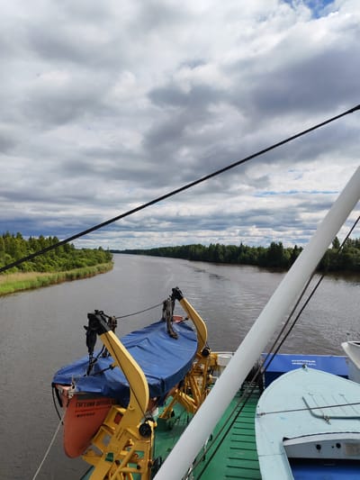 The image captures a view from a ship navigating a wide river flanked by lush green trees on both banks. The sky is overcast with dramatic, grey clouds. In the foreground, on the deck of the ship, a life raft is visible, covered by a blue tarp and mounted on a yellow davit. The name "Евгений Ороловский" and "КОТЛАС" are visible on the side of the life raft. To the right, part of another boat or vessel, painted light blue, is visible. The river appears calm, reflecting the cloudy sky. This scene likely depicts a journey along the Vytegra River, a waterway that connects Lake Beloye to Lake Vygozero in the Vologda Oblast of Russia, and is a part of the unified water system that includes the Volga-Baltic Waterway.