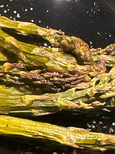 A close-up image of asparagus spears cooking in a black pan, likely in a kitchen in the city of Saint-Josse-ten-Noode, Belgium, at the coordinates provided. The asparagus spears are piled on top of each other in a layer, with some oil or other liquid visible between the spears, giving them a glossy, wet appearance. The asparagus spears are cooked to a slightly brown and tender consistency, with some charring visible. The image is in focus and shows the texture and detail of the asparagus. The asparagus spears are the subject of the image, with no other objects or people visible. The background is a blurred black surface. The asparagus spears are the only action visible in the image, and they are cooking in the pan.