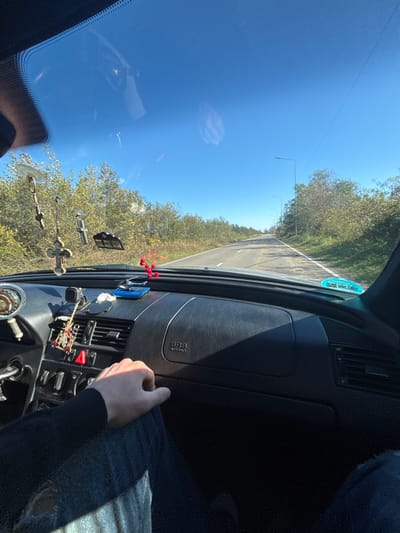 A view from inside a car, angled slightly upward. The road stretches ahead, flanked by trees under a clear blue sky. The dashboard is visible, featuring vents, controls, and an "SRS" label. Hanging ornaments dangle from the rearview mirror. A person's hand rests on the passenger side dashboard, wearing a dark sweater. The sun shines brightly, reflecting off the windshield.