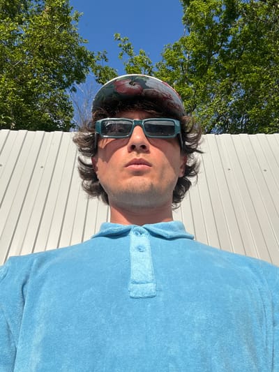 A close-up, low-angle outdoor shot captures a man wearing a light blue terry cloth polo shirt and a patterned baseball cap, facing the camera with sunglasses on. He is positioned in front of a white corrugated metal fence. Behind him, lush green trees with full foliage are visible against a clear blue sky. The scene appears to be set in Shirokaya Balka, Russia, bathed in bright sunlight, casting shadows. The man's expression is neutral, and his hair is dark and styled with some volume. The image focuses on the man's upper body and face.