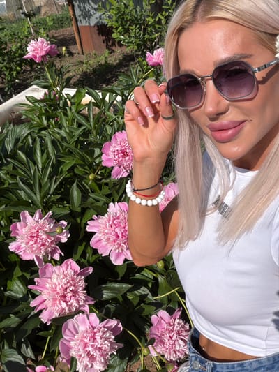 A blonde woman, wearing a white t-shirt and blue jeans, is taking a selfie outdoors. She is wearing sunglasses and has her hand near her face. Behind her, a bed of pink peonies is in bloom. The background suggests a residential area with a fence and some greenery. The image does not provide enough detail to determine the specific location within Aryshkhazda, Russia.