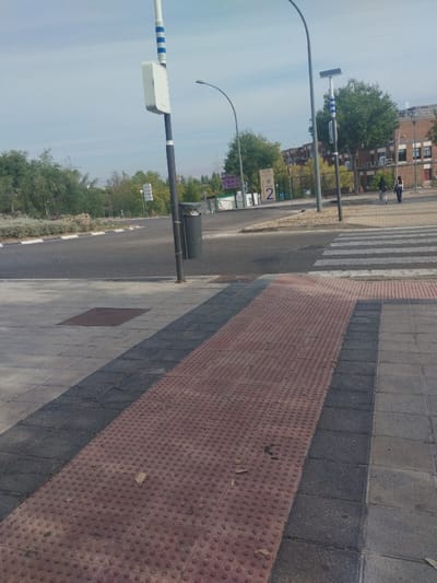 The image depicts an outdoor urban environment, specifically a street intersection or junction within the Húmera-somosaguas-prado Del Rey area of Spain, identifiable as part of a university campus due to a "UCM" sign. The time of day is daylight under a clear to partly cloudy sky.
In the immediate foreground, a tiled sidewalk features light grey and dark grey rectangular paving stones. A reddish-brown tactile paving strip with raised circular studs runs diagonally across the sidewalk from the lower left towards the mid-right. Several dried leaves are scattered on this tactile strip. A rectangular metal manhole cover is visible on the light grey paving to the left of the tactile strip.
Beyond the sidewalk, an asphalt road surface is visible. A white-striped pedestrian zebra crossing extends from the sidewalk across the road. Curbstones separate the sidewalk from the road.
Various street infrastructure elements are present:
* A tall, dark grey lamppost with blue and white stripes and a rectangular white utility box is situated on the left sidewalk. A dark grey cylindrical waste bin is attached to its base.
* A curved, dark grey lamppost spans over the road in the center.
* Another dark grey lamppost with blue and white stripes and a solar panel on top stands on the right sidewalk.
* Traffic signage includes a vertical white sign with horizontal bars on the left side of the road.
* A prominent sign displaying "UCM" (Universidad Complutense de Madrid), "LOCAL," and a large numeral "2" is visible on the right, indicating a campus entrance or area. Other indistinct purple and white signs are nearby.
The background features green trees and shrubs. Multi-story brick buildings with multiple windows are visible on the far right. Two individuals, indistinct in detail, are walking on a sidewalk near these buildings, moving away from the viewer.