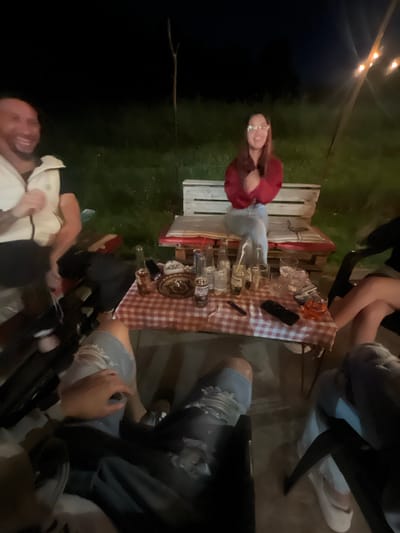 A gathering is occurring outdoors at night. A woman in glasses and a red shirt stands near a table covered with a red-and-white checkered cloth, upon which there are beer bottles and glasses. Several people are seated around the table; one individual is wearing torn jeans. String lights are visible in the background. The setting appears to be grassy. I am unable to determine the location in the city of Касапите, Bulgaria.