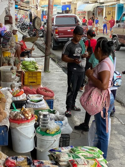 This image captures a vibrant street scene, likely in a market, in what appears to be San José de Barlovento, Venezuela. Several people interact in a crowded outdoor market setting. A man in a black Adidas tracksuit and cap holds an object, potentially money or goods, in his hands, as a woman with a pink floral backpack looks at the object. The road is lined with various goods on display, including fruits like bananas and pineapples, sacks of grain, and onions in buckets. An old-fashioned scale hangs from a metal support structure, behind which a seated man appears to be weighing goods. In the background, several motorcycles are parked. Buildings are visible on the other side of the street, and a red pickup truck parked nearby. The OCR identifies the writing "CORPORACION SUPREMO MYRIGINA" on one of the buildings.
