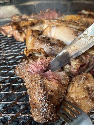 The image shows a close-up of grilled meat, likely beef, being cut with a knife and held with a fork on a metal grill. The meat is cooked to medium-rare, with a visible pink interior and browned exterior. The grill is placed on a blackened surface, possibly charcoal. The location in the city of Merlo, Argentina is not discernable from the image.
