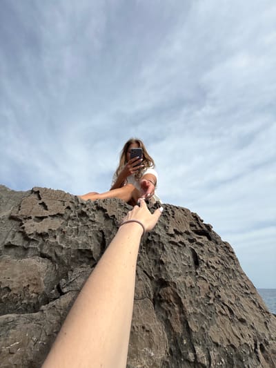 A low-angle shot shows a woman sitting on the edge of a dark rock formation under a cloudy sky. She is holding a smartphone and looking at the screen. An outstretched arm with fair skin and a bracelet is extended towards her. The location is Seoca, Montenegro.