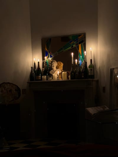 The image depicts a dimly lit interior room located in Mauléon-d'Armagnac, France, illuminated exclusively by multiple lit candles. A white-painted fireplace mantelpiece occupies the central mid-ground. Above the mantelpiece, a rectangular mirror with a stained-glass-style frame, featuring geometric patterns in blue, green, yellow, and red, reflects the candlelight and parts of the room.
Upon the mantelpiece, numerous objects are arranged:
* Approximately ten lit candles of varying heights. Several candles are inserted into the necks of dark green glass bottles, which appear to be wine or champagne bottles. At least three freestanding pillar candles or candles in holders are also present.
* Two classical-style busts are centrally positioned. The front bust is white, likely marble, depicting a female figure. Behind and slightly to its right is another bust with a warmer, possibly terracotta or aged marble, hue.
* A collection of dark glass bottles, some acting as candle holders and others standing independently, are distributed across the mantelpiece.
The walls surrounding the fireplace are white, softly illuminated by the warm, flickering candlelight, creating areas of glow and deep shadow. To the far left, on a dark surface, a large, irregular, layered object, possibly a geode or fossil, is visible. The floor in the lower foreground displays a checkered pattern of dark and lighter squares. To the far right, a dark framed item leans against the wall, and a low, dark surface, possibly a glass-topped table with an open book, is faintly discernible. No people are present, and no actions or interactions are depicted beyond the static arrangement of objects.