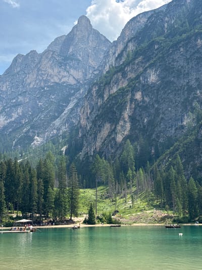 A serene lake scene is depicted, likely at Lago di Braies in Prags, Italy, with towering, rugged mountains dominating the background under a partly cloudy sky. The mountains are characterized by steep, rocky faces, with patches of green vegetation and trees clinging to their slopes. In the mid-ground, a dense forest of evergreen trees lines the shore of the luminous, turquoise lake. The water is calm, with gentle ripples visible.

On the shore, a sandy area is populated by several individuals. Some are standing and walking along the beach, while others are seated or gathered around a white umbrella providing shade. A wooden pier extends into the water on the left side of the image, with several small boats docked alongside it.

In the lake, multiple rowboats are scattered across the water. Two boats are particularly noticeable: one on the left with several people aboard, and another in the mid-right, also occupied by individuals rowing. The overall impression is one of tranquility and natural beauty, with human activity present but harmoniously integrated into the landscape.