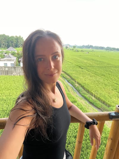 A selfie taken by a woman with brown hair and blue eyes. She is wearing a black tank top and a necklace. Her hair appears wet. She is standing on a wooden platform with a bamboo railing. Behind her is a lush green rice field and a building with a gray roof. The photo was taken in Singakerta, Indonesia. A black scrunchie adorns her left wrist as she rests it on the railing. A small black device is attached to the railing.