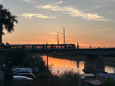 In Mannheim, Germany, a train crosses a bridge over a body of water during sunset. The sky is painted with hues of orange and yellow, with soft clouds scattered across it. The train, silhouetted against the vibrant sky, is traveling from left to right. Below the bridge, the water reflects the warm colors of the sunset, creating a shimmering surface. On the foreground, parked cars and greenery are visible. The overall scene depicts a tranquil urban landscape bathed in the warm glow of the setting sun.