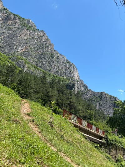 The image depicts a steep, grassy hillside in Zgorigrad, Bulgaria. A narrow dirt path winds its way up the slope, bordered by lush green grass. In the middle ground, a small bridge with red and white railings spans a depression in the terrain. The bridge appears to be constructed of concrete and stone. Behind the bridge and the hillside, a massive, rugged mountain looms, its rocky face streaked with grey and dotted with patches of green vegetation. The sky above is a clear, vibrant blue. In the distance, to the right of the bridge, a road sign with Cyrillic lettering and an arrow pointing to the left is visible, indicating "Academia" and "Ledenika."