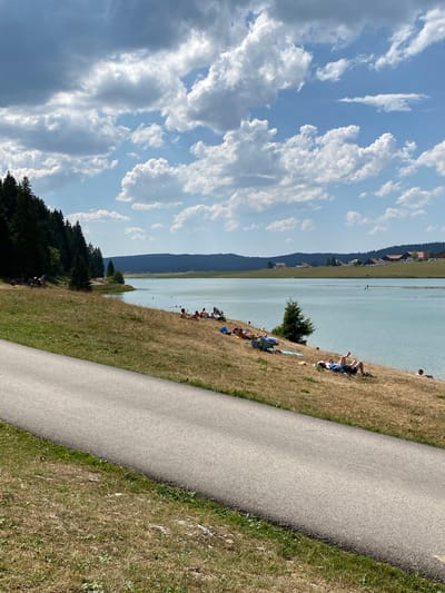 This is an eye-level, medium shot of a lake with a group of people sunbathing on its grassy, dry bank in La Brévine, Switzerland. The sky is partly cloudy with dramatic white clouds against a blue background. A paved path runs diagonally across the foreground from the bottom left to the middle right of the frame. On the grassy bank, several individuals are lying down, sunbathing, or sitting. Some are on towels or blankets. One person is visible in the water. In the distance, across the lake, a line of trees forms the horizon, with houses dotting the landscape behind them. The trees on the left side of the frame are dark green and dense. The water is a pale blue-green, and the grass on the bank is dry and golden, suggesting a warm season. The overall atmosphere is one of a relaxed, sunny day at a lake.