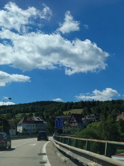 The image captures a view from a vehicle on a road approaching a town nestled amongst forested hills. The sky is a bright blue with scattered white clouds. The road curves to the right, with a metal guardrail on the right side. Several cars are visible on the road, including a black van in the foreground and a dark-colored car on the left.
In the background, a cluster of buildings forms a small town. One prominent building features a traditional half-timbered architectural style with a red roof. Adjacent to it is a larger, light-colored building with multiple windows. The town is surrounded by lush green trees that extend up the hillsides.
A blue traffic sign with white arrows indicating lane direction is visible on the right side of the road. A street lamp is also present on the left side of the road, near the crest of the hill. The overall scene depicts a scenic drive through a rural European landscape, leading into a village or small town. There is no clear indication that this location is specifically Vimperk, Czechia, as there are no identifiable landmarks or text to confirm the location.