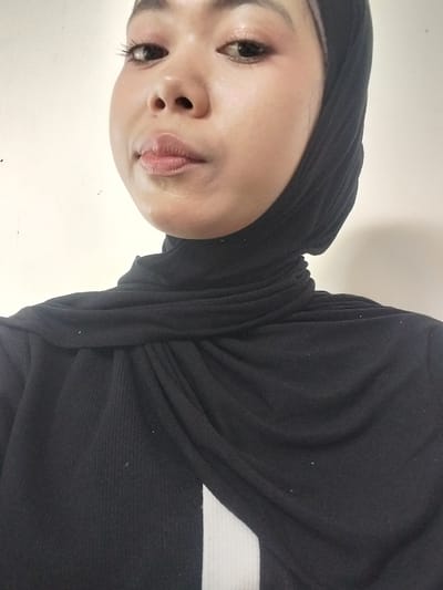 A close-up, waist-up selfie shows a young woman wearing a black hijab and a black ribbed top. She is looking towards the camera with her head slightly tilted to the right. Her makeup includes noticeable eyeliner and lipstick. The background is a plain, off-white wall. The image appears to be taken indoors, likely in a domestic setting. There is no indication of the city of Jeppee, Indonesia, in the image or its context.