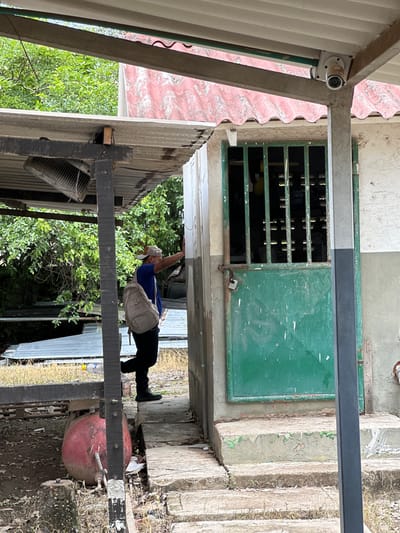 The image shows a man with a backpack standing in front of a green metal door with a padlock in Juan Mina, Colombia. He is wearing a blue t-shirt and black pants, and a baseball cap. The door is on a small, light-colored building with a green-grated window above it. The building has a pink corrugated metal roof. The man appears to be reaching towards the door. To the left of the building, there is a covered area with a corrugated metal roof and what appears to be a broken ventilation unit. In the foreground, on the ground to the left, is a large, round, red tank with pipes. The environment is overgrown with vegetation, and there are scattered debris and metal sheets on the ground. A security camera is visible mounted on the underside of the overhang above the door.