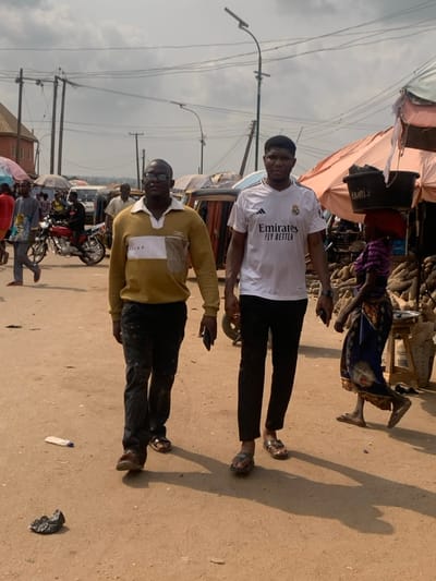 The scene depicts a bustling open-air market in Otuocha, Nigeria, under a bright, overcast sky. Two adult males occupy the foreground, walking towards the viewer's right. The individual on the left wears a gold-yellow long-sleeved polo shirt with a white horizontal stripe featuring "VOLUF" text, dark trousers, and brown sandals; he holds a dark smartphone in his right hand. The individual on the right wears a white short-sleeved t-shirt with an Adidas logo, Real Madrid crest, and 'Emirates FLY BETTER' text, black trousers, and gold-brown sandals; he holds a dark smartphone in his left hand.
The ground is packed earth, with a clear plastic bottle and a crumpled dark plastic bag visible as litter in the foreground. In the mid-ground on the right, an adult female with a red head covering walks past market stalls, balancing two dark buckets marked 'EMEFIA 2' on her head. These stalls are covered by tarpaulin canopies (one orange, one blue) and display numerous piles of root vegetables, likely yams, alongside sacks and a silver tray on a wooden stool.
In the left mid-ground, a red motorcycle is parked, and various individuals are seen walking. The background features several auto-rickshaws and a white van. Numerous utility poles, overhead power lines, and two solar-powered streetlights dominate the upper section of the image. Distant buildings and a hazy horizon are visible. Shadows are cast by the foreground figures, indicating a strong light source.