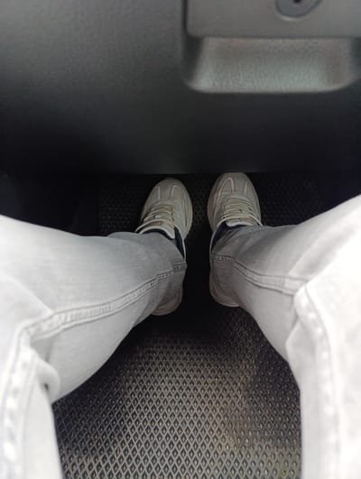 A first-person perspective shot inside the footwell of a vehicle shows a person wearing light-wash jeans and gray running shoes with white soles. The feet are resting on a black rubber floor mat with a diamond pattern. The lower portion of the dashboard is visible at the top of the frame. The location is likely Володарский, Russia.