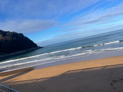The image captures a coastal scene in Zarautz, Spain, from an elevated vantage point. The foreground features a wide expanse of light brown sand, stretching from the bottom of the frame towards the ocean. Distinct, elongated shadows from an unseen structure in the immediate foreground are cast diagonally across the sand, extending from the lower right to the mid-left. Faint footprints are discernible on the sand.

The midground is dominated by the Atlantic Ocean, exhibiting varying shades of blue and green. Several lines of white-capped waves are actively breaking onto the shore. To the left, a dark, densely tree-covered headland or hill extends into the water, forming a natural barrier. A light-colored structure, possibly a walkway or sea wall, is visible at the base of this landmass where it meets the water.

The sky above is clear and bright blue, interspersed with wispy white clouds. A metallic railing, likely part of a staircase or viewing platform, is partially visible in the bottom-left corner of the frame. No individuals, animals, or explicit human activities are present within the scene.