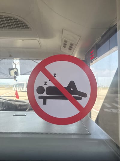 A circular "No Sleeping" prohibition sign, featuring a black pictogram of a recumbent figure with two "Z"s on a white background intersected by a red circle and diagonal slash, is affixed to a transparent window. The scene is observed from within a vehicle or structure at an airport on a clear day in Hòa Cường Ward, Da Nang, Vietnam. Through the window, a tarmac is visible, displaying a distant white ground support vehicle or bus and a red flag. Interior elements include a light grey overhead panel with a ventilation grille and a rectangular light fixture containing six small circular lights, a rearview mirror partially reflecting an external scene, and a light grey window ledge. Partial red LED lights are visible on the far right, outside the window.