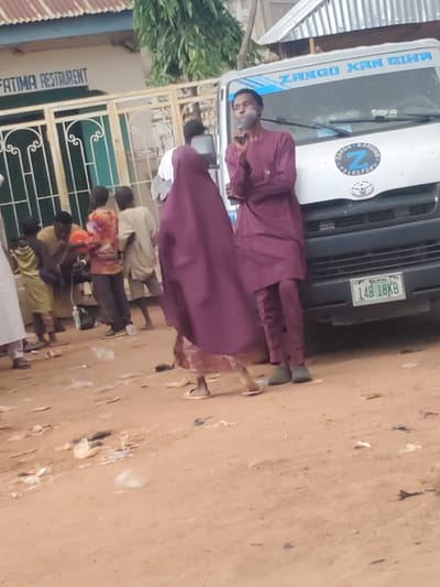 The image depicts an outdoor scene in Funtua, Nigeria, likely in front of a business. A white van with blue lettering that reads "ZANGO KAN GIHA" and a circular emblem with a "Z" is parked on a dirt or gravel surface. License plate "14B-18KB" is visible on the front of the van. To the left of the van, a building with a corrugated metal roof and a sign that partially reads "FATIMA RESTAURANT" is visible. A decorative metal gate is in front of the restaurant.
Several individuals are present in the scene. In the foreground, a person in a maroon outfit, including a long tunic and pants, stands facing towards the van, holding what appears to be a cigarette. Another person in a similar maroon garment is partially visible to the left, also appearing to be in motion. Further back, several other people are gathered. One individual in a colorful patterned shirt and dark pants is holding a red spherical object. Another person in a light-colored tunic and shorts stands nearby. A person in a yellow outfit is visible to the left, and a person in a white garment is partially seen in the far left. The ground is sandy and littered with debris, including plastic wrappers and discarded items. The overall impression is of a public space with moderate activity.