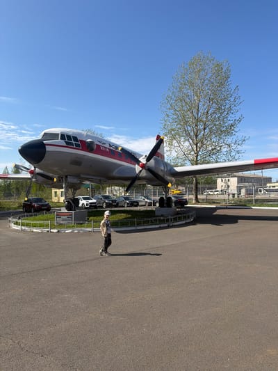 An outdoor scene shows an Ilyushin Il-14 airplane on display at the Arkhangelsk Regional Aviation Museum in Arkhangelsk, Russia. The plane is gray with red and white markings. Its propellers are black with yellow tips. A young person stands near the plane, casting a shadow on the asphalt. Behind the plane is a row of parked cars and buildings in the background. A tree with budding leaves stands next to the plane. The sky is blue with some scattered clouds.