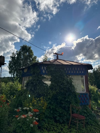 This image depicts a sun-drenched outdoor scene in Bolshiye Turyshi, Russia, featuring a rustic gazebo heavily overgrown with green vines. The gazebo has a weathered brown roof and blue lattice trim with red diamond accents. A decorative airplane weather vane sits atop the roof. In the foreground, a cluster of orange and white flowers bloom amidst lush green foliage. A solitary wooden chair with a red cushion is positioned to the right of the gazebo, partially obscured by the greenery. The background consists of dense trees and a bright blue sky with scattered white clouds, with the sun prominently visible, creating a lens flare. A power line runs diagonally across the upper portion of the frame. The overall impression is one of a slightly overgrown, rural, or dacha-like setting.